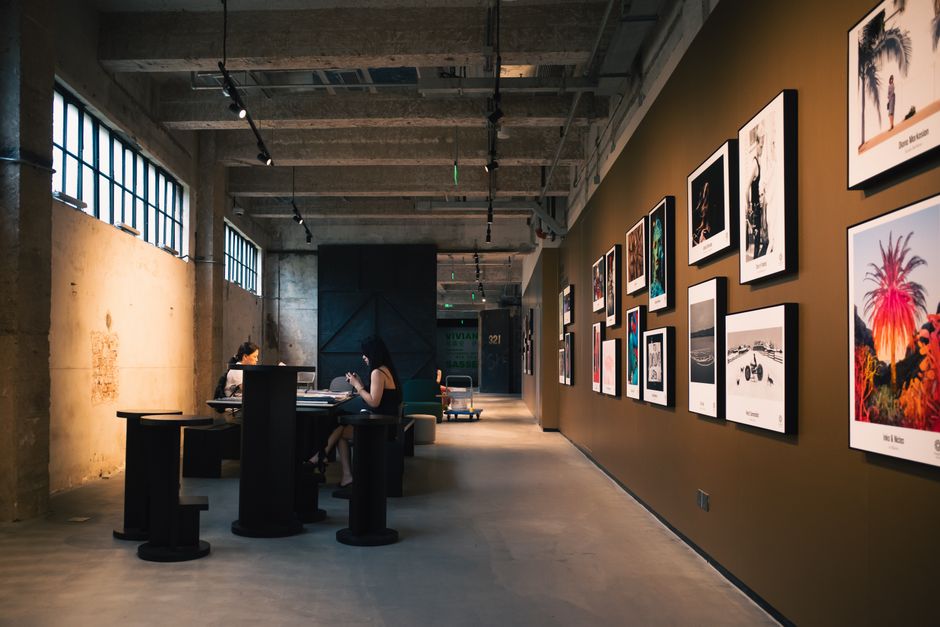 Art gallery hallway with framed artworks and visitors seated inside, under warm lighting.