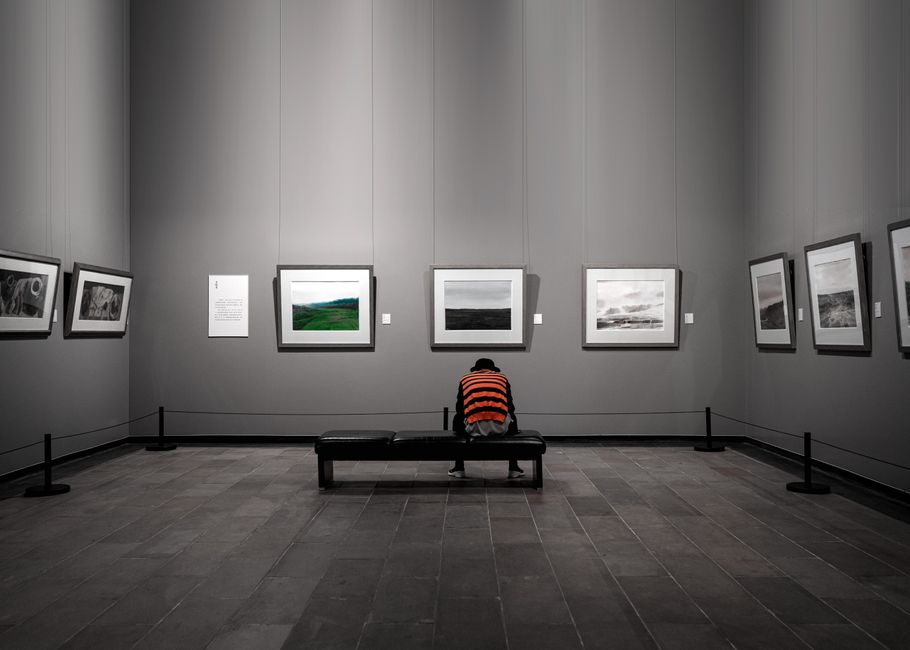 A solitary figure sitting in a contemporary art gallery surrounded by framed artworks.